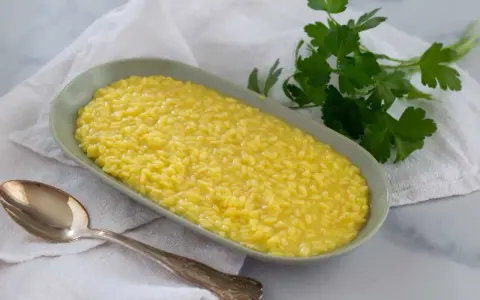 Un cremoso risotto allo zafferano in un piatto ovale, con un cucchiaio d'argento e un rametto di prezzemolo fresco.