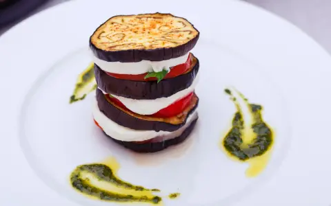 Parmigiana di melanzane a strati, con mozzarella, pomodoro e basilico, guarnita con pesto su piatto bianco.