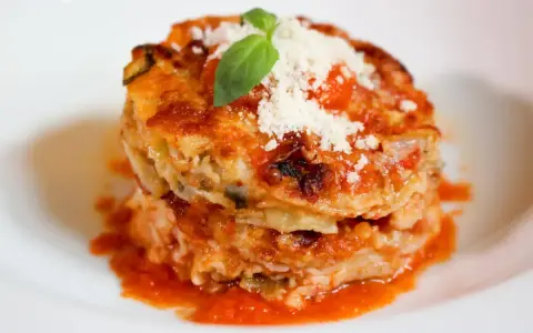 Parmigiana di alici, un piatto stratificato con salsa di pomodoro, formaggio e basilico fresco, servito su un piatto bianco.