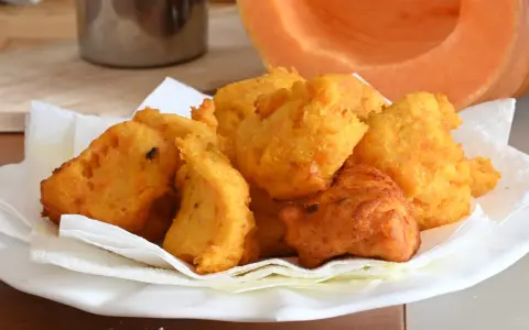 Frittelle di zucca dorate e croccanti, servite su carta assorbente, con una fetta di zucca sullo sfondo.