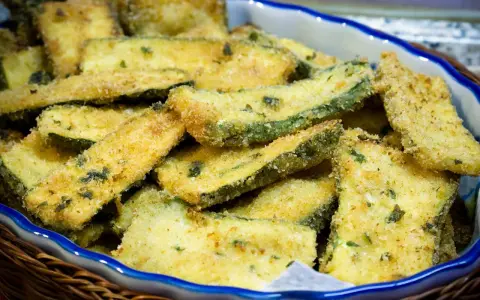Fette di zucchine gratinate in padella, dorate e croccanti, servite in una ciotola blu bordata di bianco.