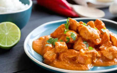Spezzatino di pollo cremoso e speziato, guarnito con prezzemolo fresco, servito su un piatto blu con riso bianco e spicchio di lime.