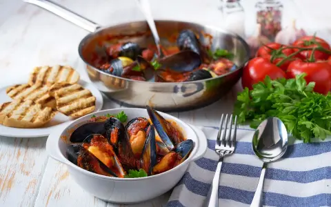 Cozze alla tarantina fumanti in una ciotola bianca, accompagnate da pane grigliato e ingredienti freschi.