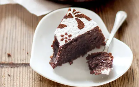 Una fetta di torta caprese ricetta originale napoletana, cosparsa di zucchero a velo e cacao, su un piattino bianco a forma di cuore.