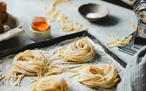 Nidi di pasta fatta in casa su un vassoio infarinato, con uova e una macchina per la pasta sullo sfondo.