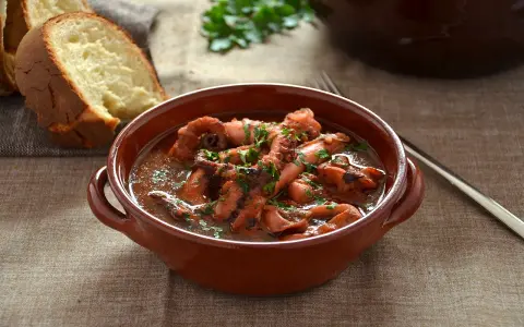 Un saporito polpo alla pignata, cotto lentamente in umido, servito in una ciotola di terracotta con pane fresco a lato.
