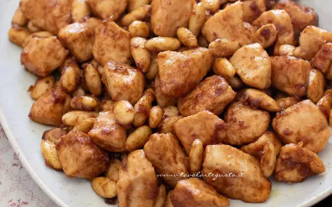 Close-up of bite-sized chicken pieces and almonds coated in a glossy brown sauce on a white plate.
