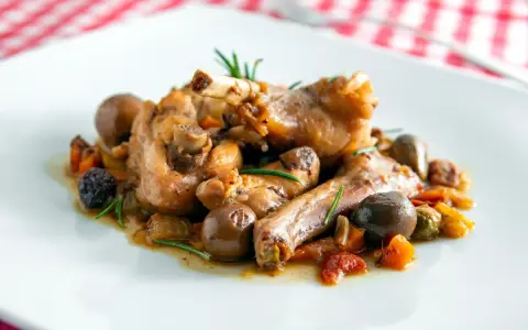 Coniglio alla cacciatora ricetta della nonna: succulenti pezzi di coniglio con olive, pomodorini e rosmarino su un piatto bianco.