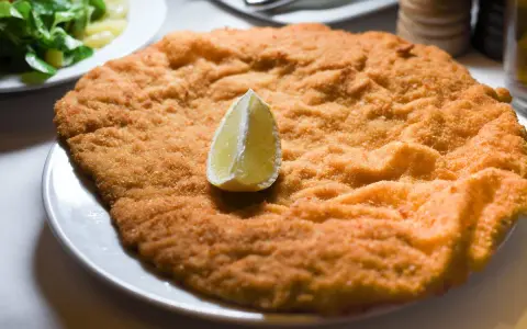 Una cotoletta dorata, grande come un'orecchia di elefante, servita con una fetta di limone e un'insalata fresca.