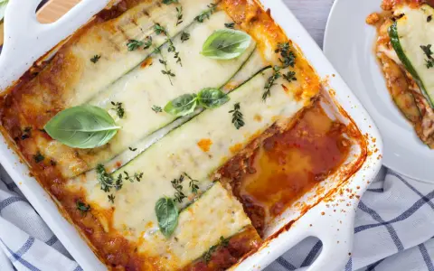 Lasagne di zucchine al forno, dorate e filanti, guarnite con basilico e timo. Una porzione è già servita su un piatto bianco.