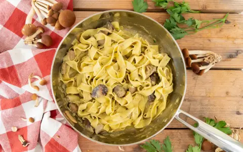Tagliatelle ai funghi cremose in padella, con funghi freschi e prezzemolo su fondo di legno.