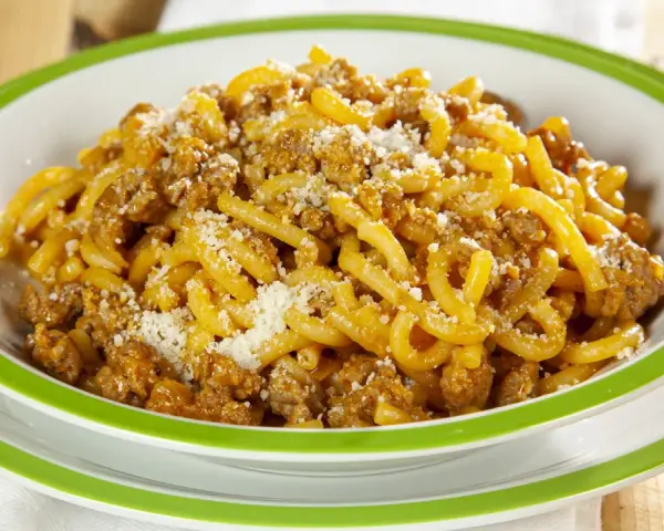 Piatto di gramigna alla salsiccia, condita con ragù e parmigiano grattugiato, servita in una ciotola con bordo verde.