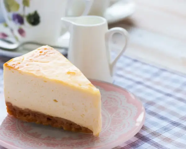 Una fetta di cheesecake con mascarpone, cremosa e invitante, su un piattino rosa. Sullo sfondo, una tazza da tè e una lattiera.