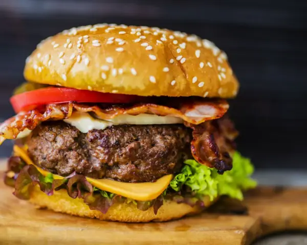 Hamburger fatti in casa con bacon croccante, formaggio filante, pomodoro fresco e lattuga su un tagliere di legno.