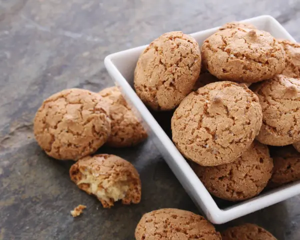 Un mucchio di croccanti biscotti alle mandorle in una ciotola bianca, alcuni sparsi su una superficie grigia.