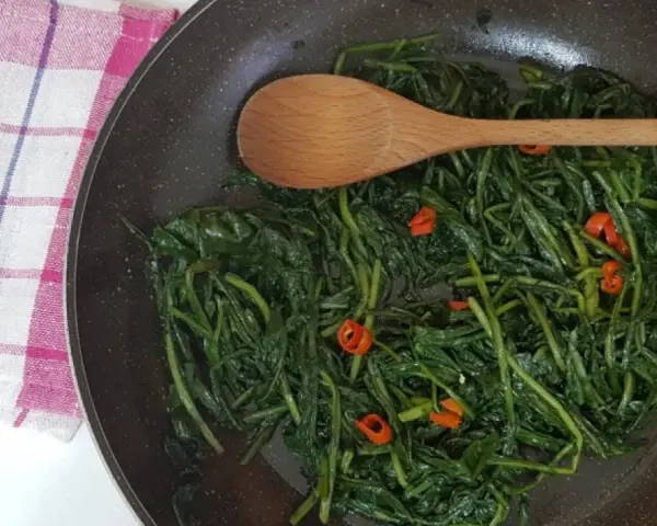 Cicoria in padella con peperoncino, un contorno saporito e salutare. Un cucchiaio di legno mescola le foglie verdi.