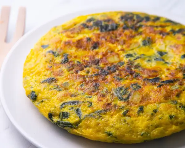 Frittata di spinaci dorata e invitante su un piatto bianco, con un utensile in legno sullo sfondo.