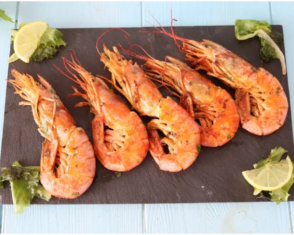 Cinque succulenti gamberoni alla griglia, serviti su una pietra scura con spicchi di limone e insalata fresca.