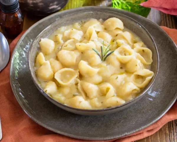 Una ciotola di minestra di patate cremosa con pasta a conchiglia, guarnita con rosmarino. Un piatto confortante e saporito.