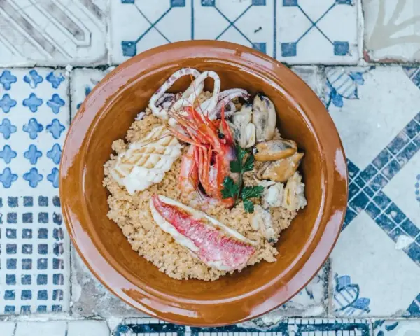 Un invitante cous cous di pesce con gamberi, calamari, cozze e pesce rosso, guarnito con prezzemolo, servito in una ciotola di terracotta su un piatto di maioliche.