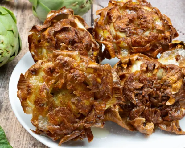 Carciofi alla giudia dorati e croccanti su un piatto bianco, con carciofi freschi sullo sfondo.