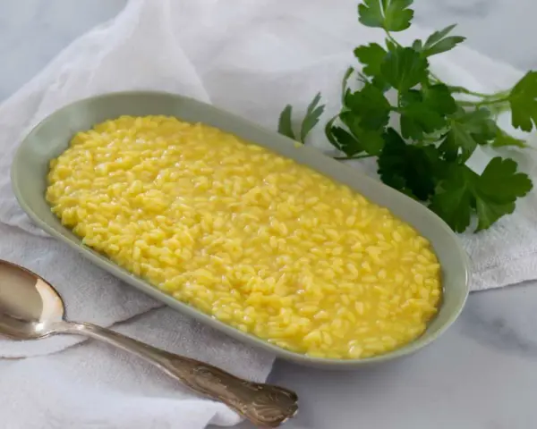 Un cremoso risotto allo zafferano in un piatto ovale, con un cucchiaio d'argento e un rametto di prezzemolo fresco.