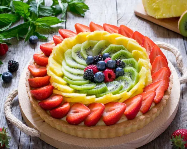 Una colorata crostata di frutta, con fragole, kiwi, ananas e frutti di bosco, su un tavolo di legno rustico.