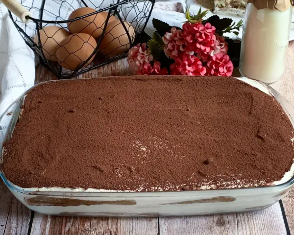 Un invitante tiramisù con panna, cosparso di cacao, pronto per essere gustato. Ingredienti freschi come uova e latte completano la scena.