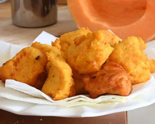 Frittelle di zucca dorate e croccanti, servite su carta assorbente, con una fetta di zucca sullo sfondo.