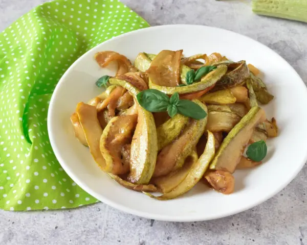 Zucchine in agrodolce, un piatto colorato e saporito, guarnito con basilico fresco su un piatto bianco, accanto a un tovagliolo a pois verdi.
