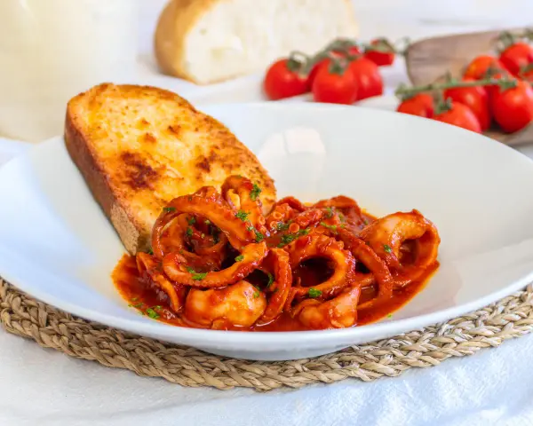 Un piatto invitante di moscardini in umido, con tentacoli succulenti in salsa rossa, accompagnati da una fetta di pane tostato dorato.
