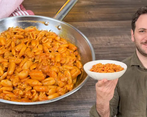 Un uomo sorride mostrando una ciotola di gnocchetti alla campidanese, accanto a una padella piena dello stesso piatto.
