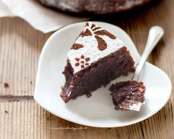 Una fetta di torta caprese ricetta originale napoletana, cosparsa di zucchero a velo e cacao, su un piattino bianco a forma di cuore.