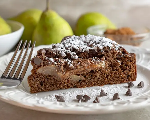 Una fetta di torta pere e cioccolato, cosparsa di zucchero a velo e gocce di cioccolato, con pere caramellate all'interno.