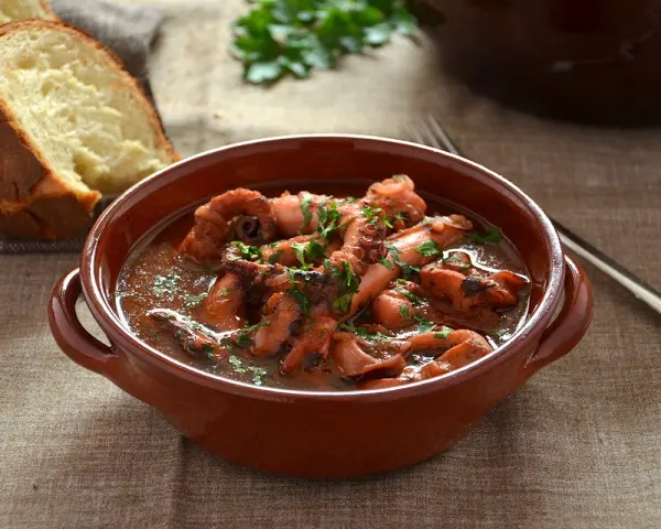 Un saporito polpo alla pignata, cotto lentamente in umido, servito in una ciotola di terracotta con pane fresco a lato.