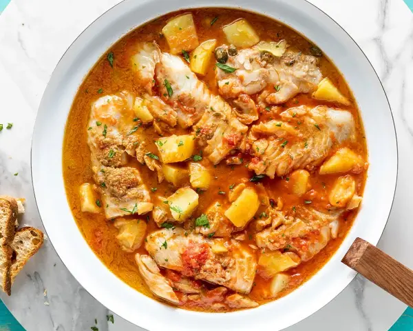 Un saporito merluzzo in umido con patate e pomodoro, guarnito con prezzemolo fresco e accompagnato da pane tostato.