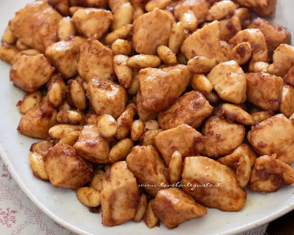 Close-up of bite-sized chicken pieces and almonds coated in a glossy brown sauce on a white plate.