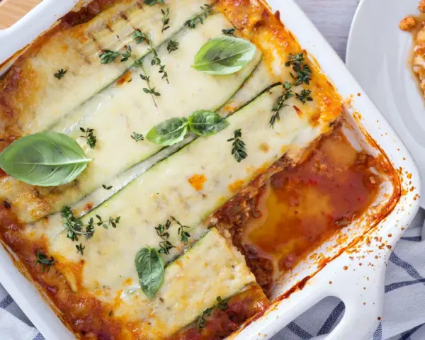 Lasagne di zucchine al forno, dorate e filanti, guarnite con basilico e timo. Una porzione è già servita su un piatto bianco.