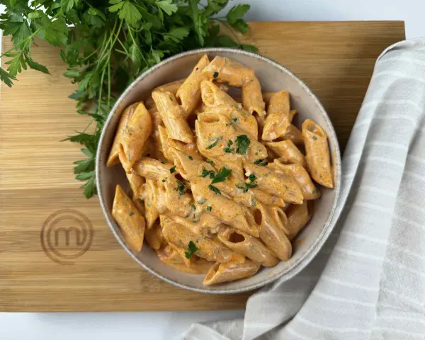 Penne alla vodka cremose e invitanti, guarnite con prezzemolo fresco, su un tagliere di legno con il logo di MasterChef.