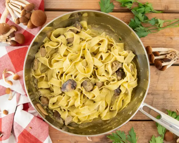 Tagliatelle ai funghi cremose in padella, con funghi freschi e prezzemolo su fondo di legno.