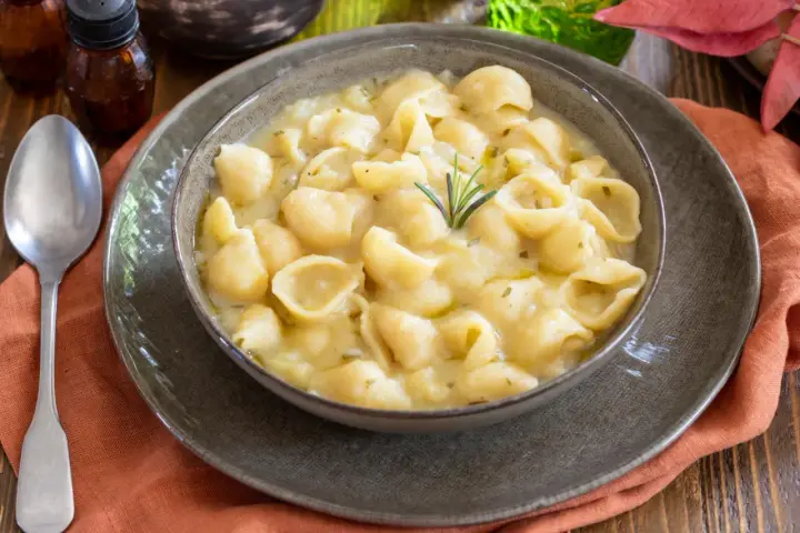 Una ciotola di minestra di patate cremosa con pasta a conchiglia, guarnita con rosmarino. Un piatto confortante e saporito.