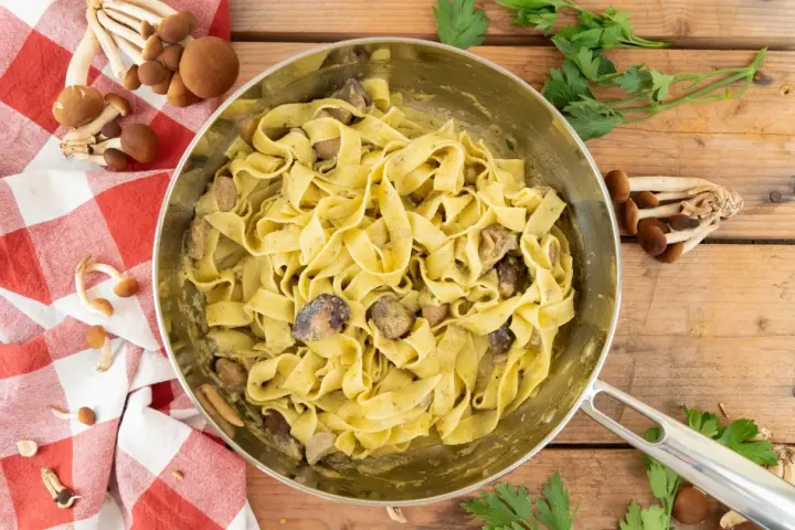 Tagliatelle ai funghi cremose in padella, con funghi freschi e prezzemolo su fondo di legno.