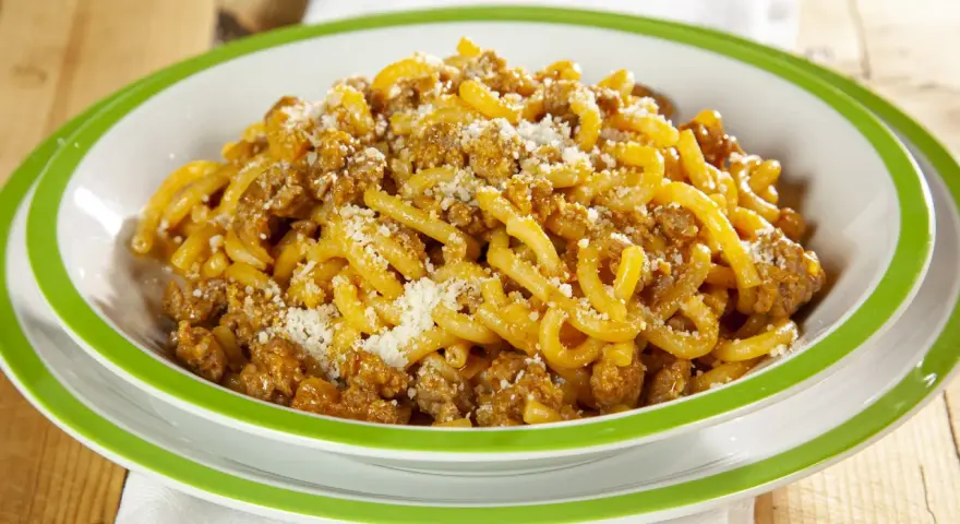 Piatto di gramigna alla salsiccia, condita con ragù e parmigiano grattugiato, servita in una ciotola con bordo verde.