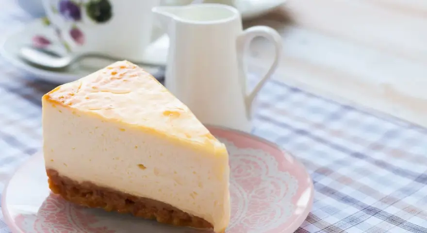 Una fetta di cheesecake con mascarpone, cremosa e invitante, su un piattino rosa. Sullo sfondo, una tazza da tè e una lattiera.