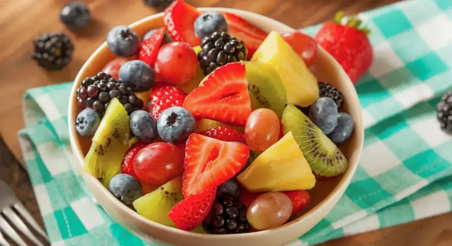 Una colorata macedonia di frutta con fragole, mirtilli, kiwi, ananas e uva, servita in una ciotola su una tovaglia a quadretti verdi.
