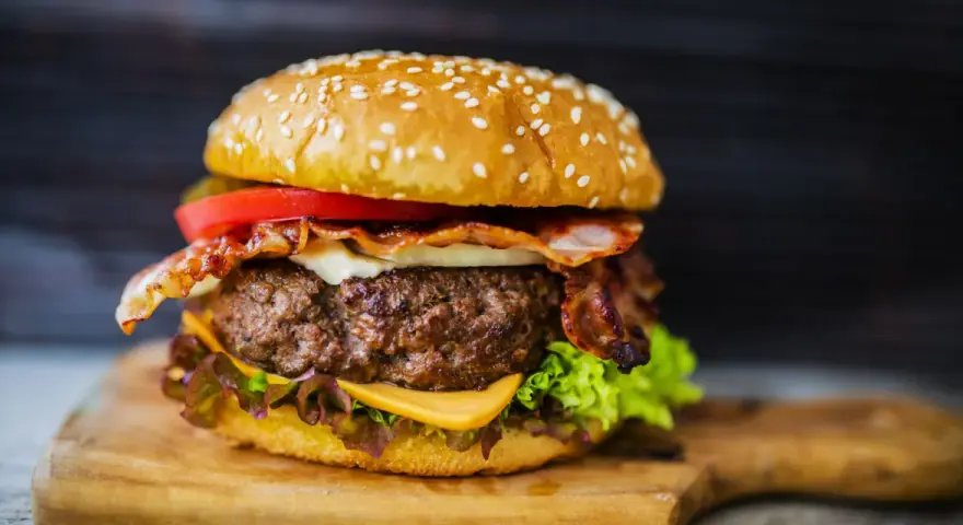 Hamburger fatti in casa con bacon croccante, formaggio filante, pomodoro fresco e lattuga su un tagliere di legno.