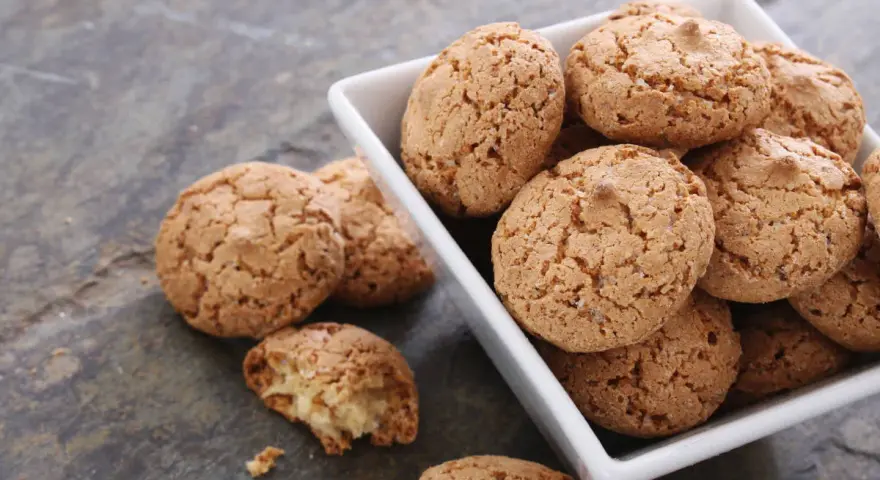 Un mucchio di croccanti biscotti alle mandorle in una ciotola bianca, alcuni sparsi su una superficie grigia.