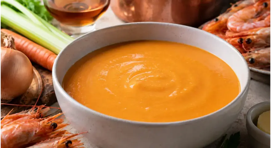 Una ciotola di cremosa bisque di gamberi, con gamberi freschi, carote, sedano e cipolla sullo sfondo.