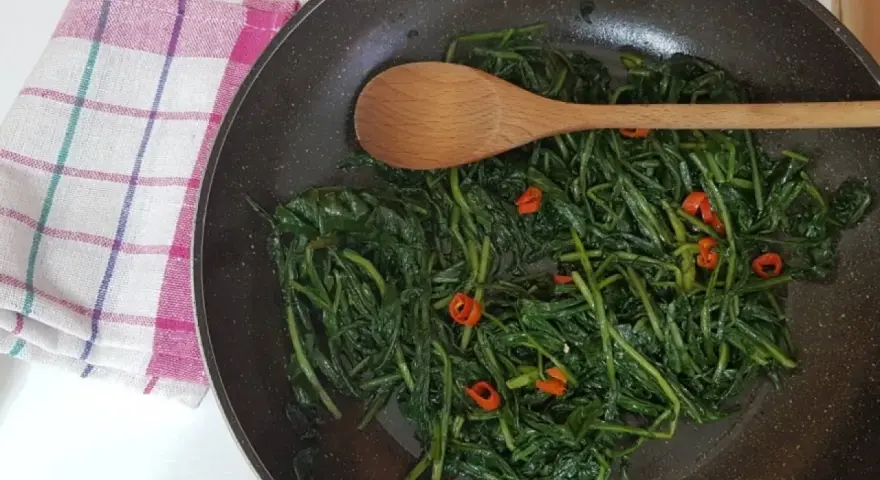 Cicoria in padella con peperoncino, un contorno saporito e salutare. Un cucchiaio di legno mescola le foglie verdi.