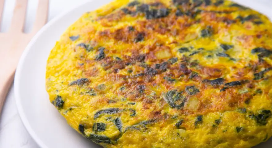 Frittata di spinaci dorata e invitante su un piatto bianco, con un utensile in legno sullo sfondo.